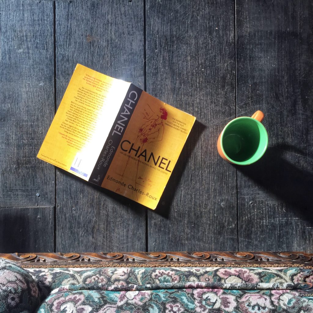 Photo of a Chanel biography book face down on a wooden floor beside an empty cup of coffee.
