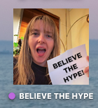 Photo of a woman with a cheesy grin holding paper that says believe the hype