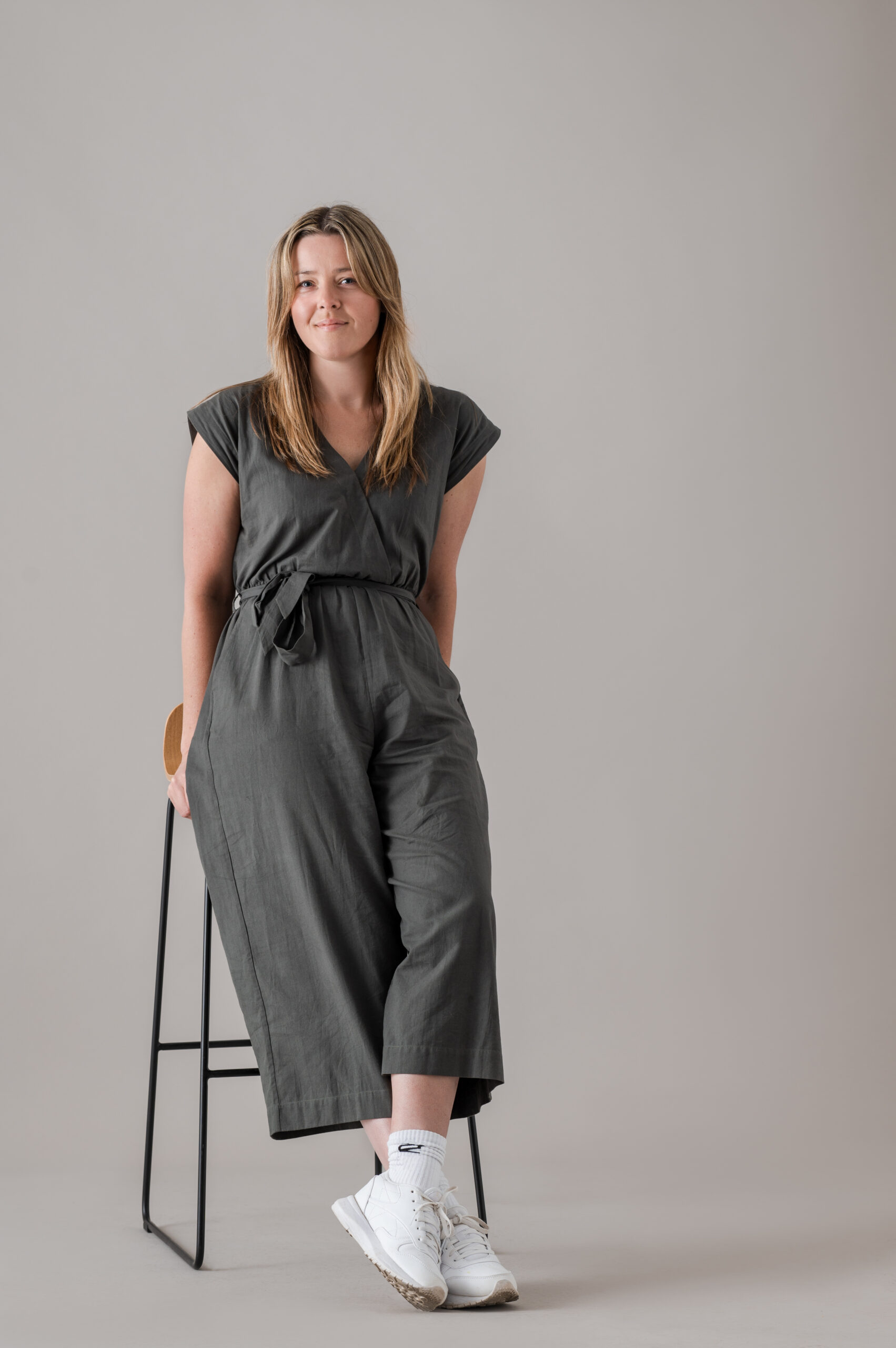 Photo of a woman wearing a khaki jumpsuit and sneakers, leaning on a stool and slightly smiling